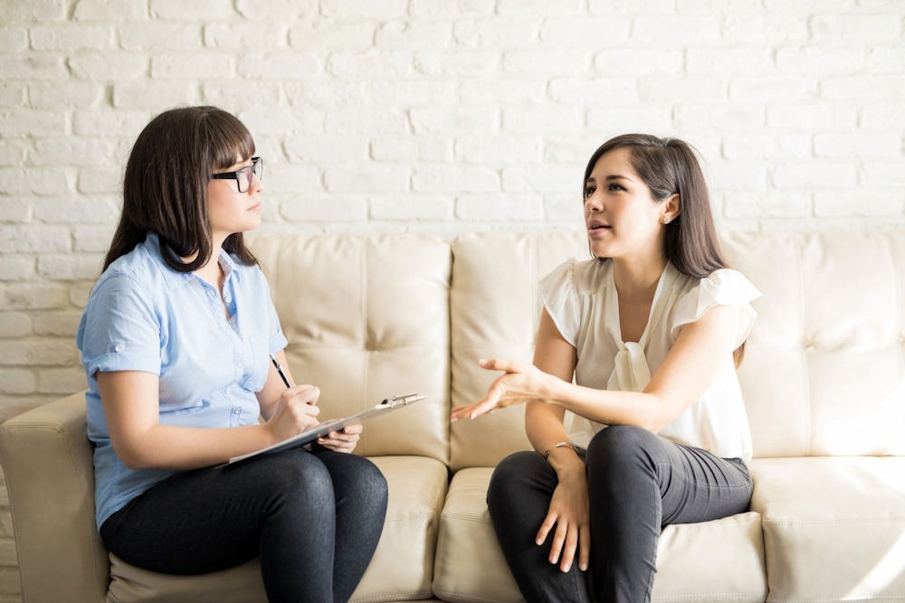 two women sitting on couch having discussion while one records on a clipboard