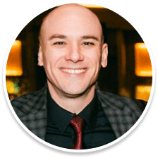 Smiling bald man wearing a dark checkered jacket, black shirt, and red tie, posing in a warmly lit indoor setting—an inspiring presence at the Mission Viejo rehab program.